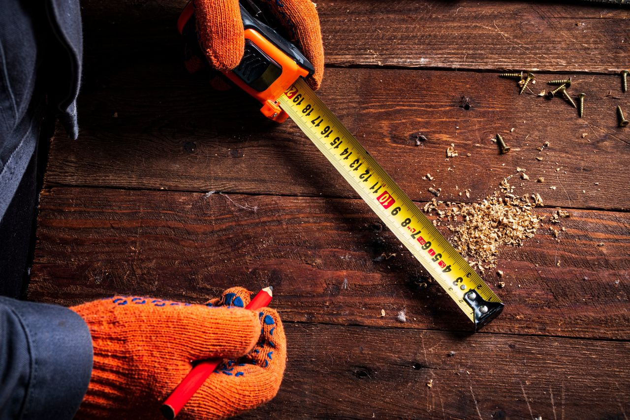 gallery-3 Close-up of gloved hands using a tape measure on wooden surface for precise carpentry.