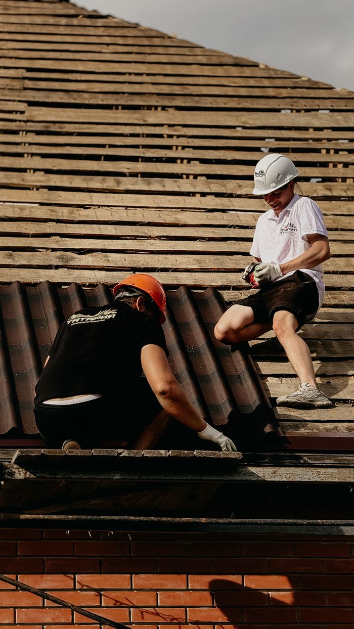 gallery-1 Two construction workers in hard hats repair roof tiles under sunny conditions. Ideal for industrial themes.