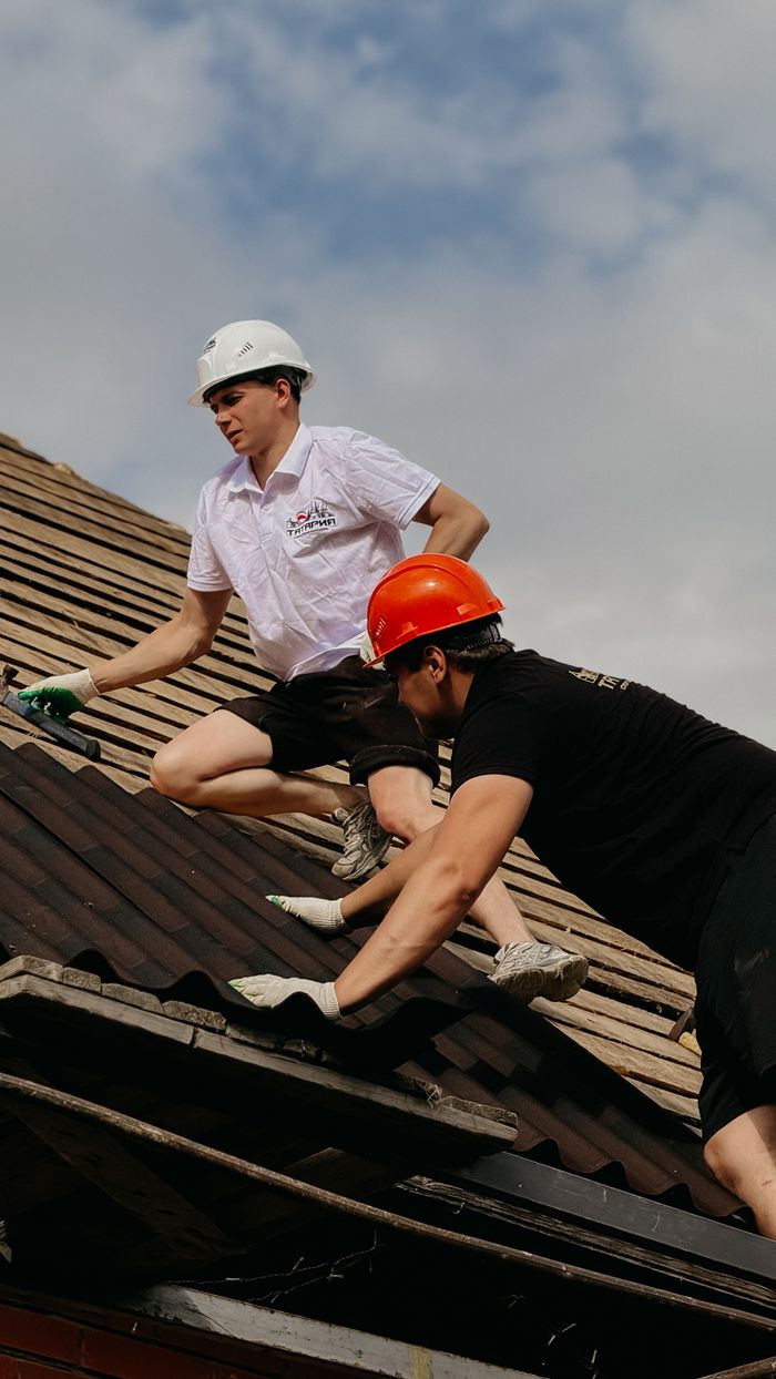 why-choose-us Two construction workers in safety helmets repairing a roof under clear skies.