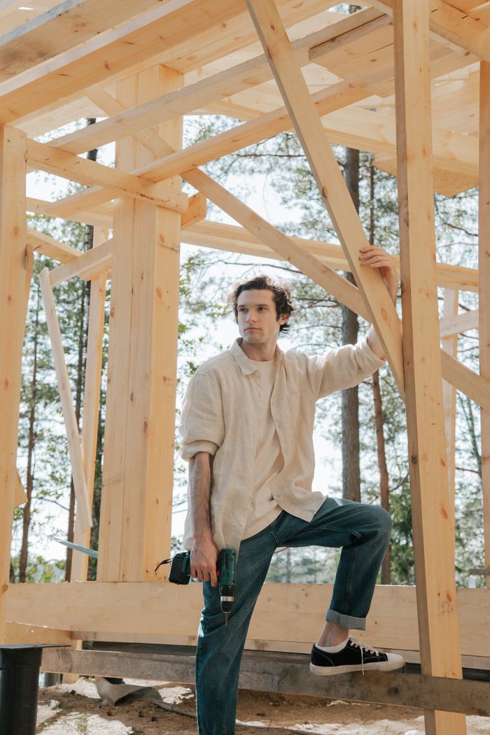 gallery-5 Man holding drill at construction site with wooden beams around, symbolizing craftsmanship and building.